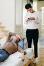 A Man in White Sweater Holding a Phone while Standing Near His Partner Sleeping on the Couch