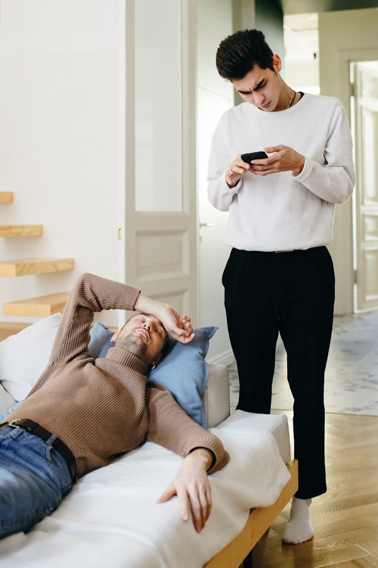 A Man In White Sweater Holding A Phone While Standing Near His Partner Sleeping On The Couch