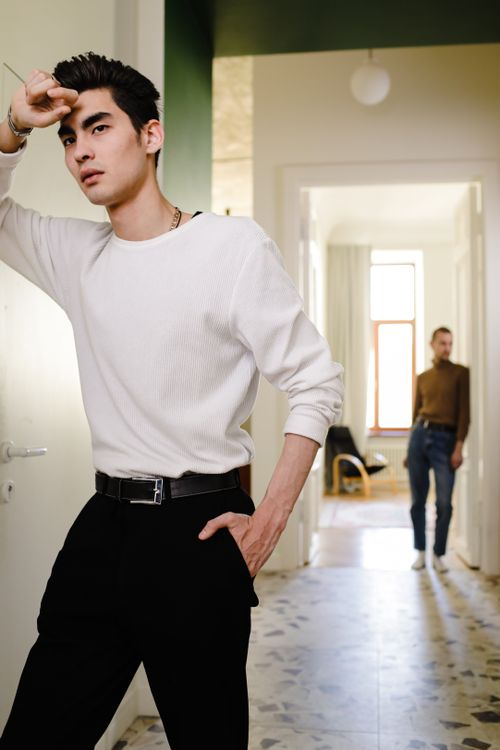 Free Young serious male model wearing white sweater and black trousers leaning on one hand and keeping hand in pocket posing in light modern studio Stock Photo