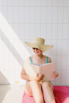 Smiling woman in swimwear reading a book while sitting in the sunshine with a straw hat.
