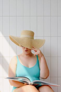 Woman in a turquoise swimsuit reading a book indoors, wearing a wide-brimmed sun hat.