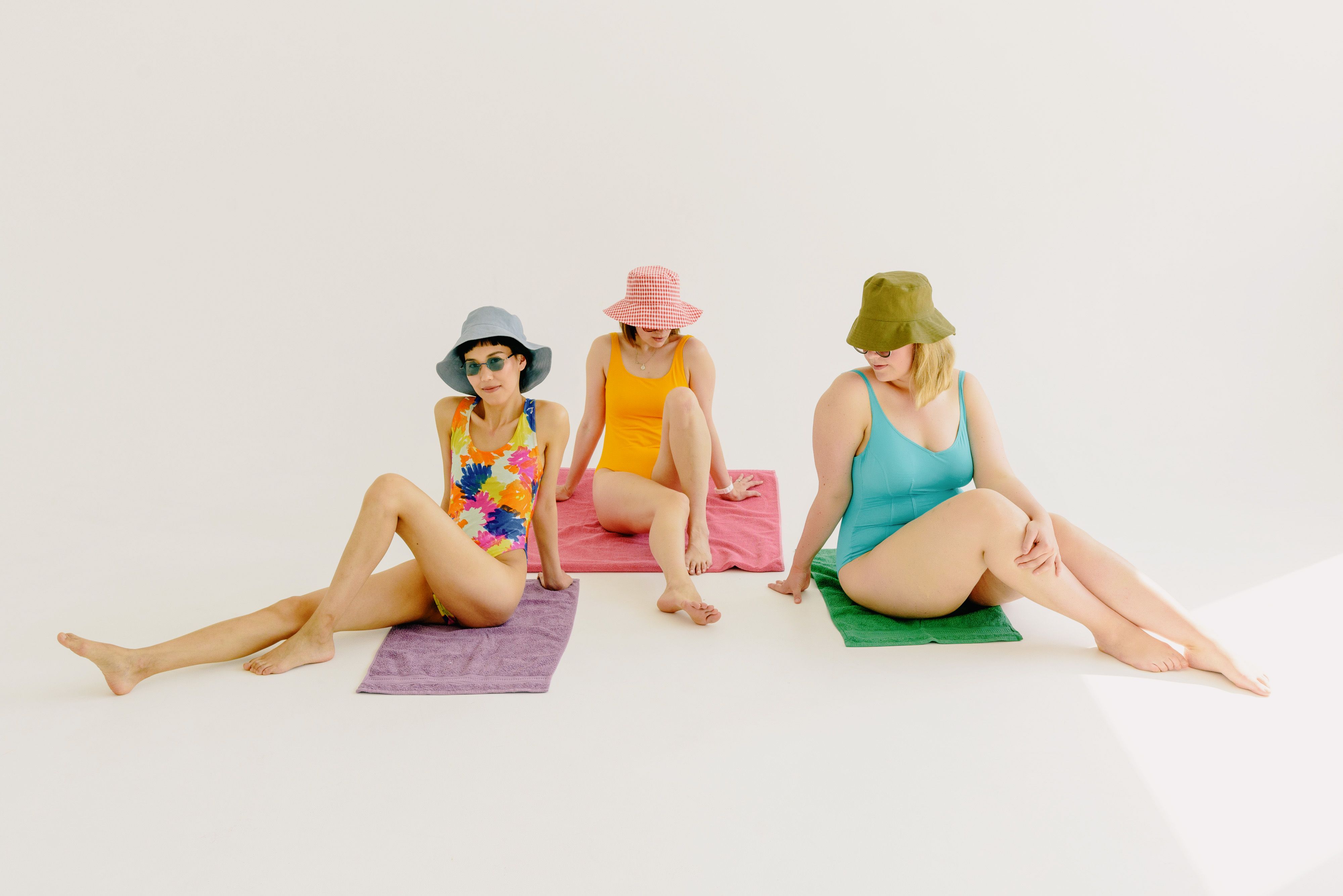 Three women in vibrant swimsuits and bucket hats sit on towels in a bright, minimalist studio setup.