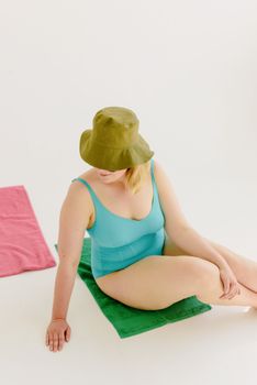 Young woman wearing a green hat and turquoise swimsuit sitting on a towel in a minimalistic setting.
