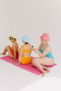 Three women in vibrant swimsuits and hats sitting on a towel indoors, enjoying a summer vibe.