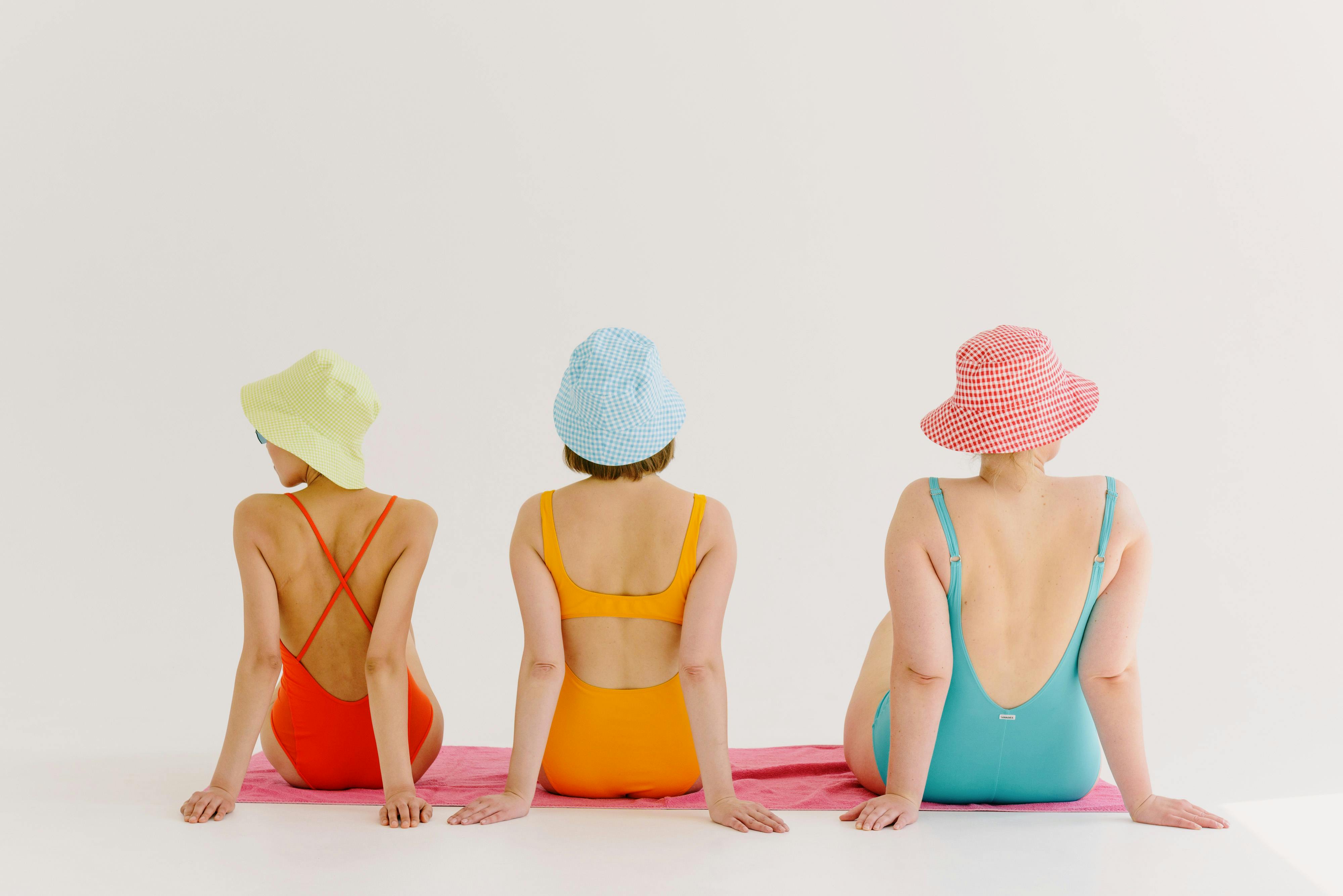 Three women in vibrant swimsuits and bucket hats sitting on a towel, back view.
