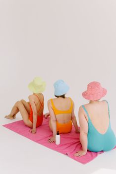 Three women in colorful swimwear and bucket hats sitting on a pink towel, enjoying a sunny day.