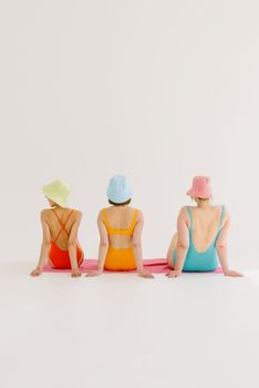 Three women in colorful swimsuits and bucket hats sitting on towels in a studio setting.