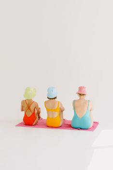 Three women in colorful swimwear and hats sit on a vibrant pink towel, showcasing a playful summer vibe.