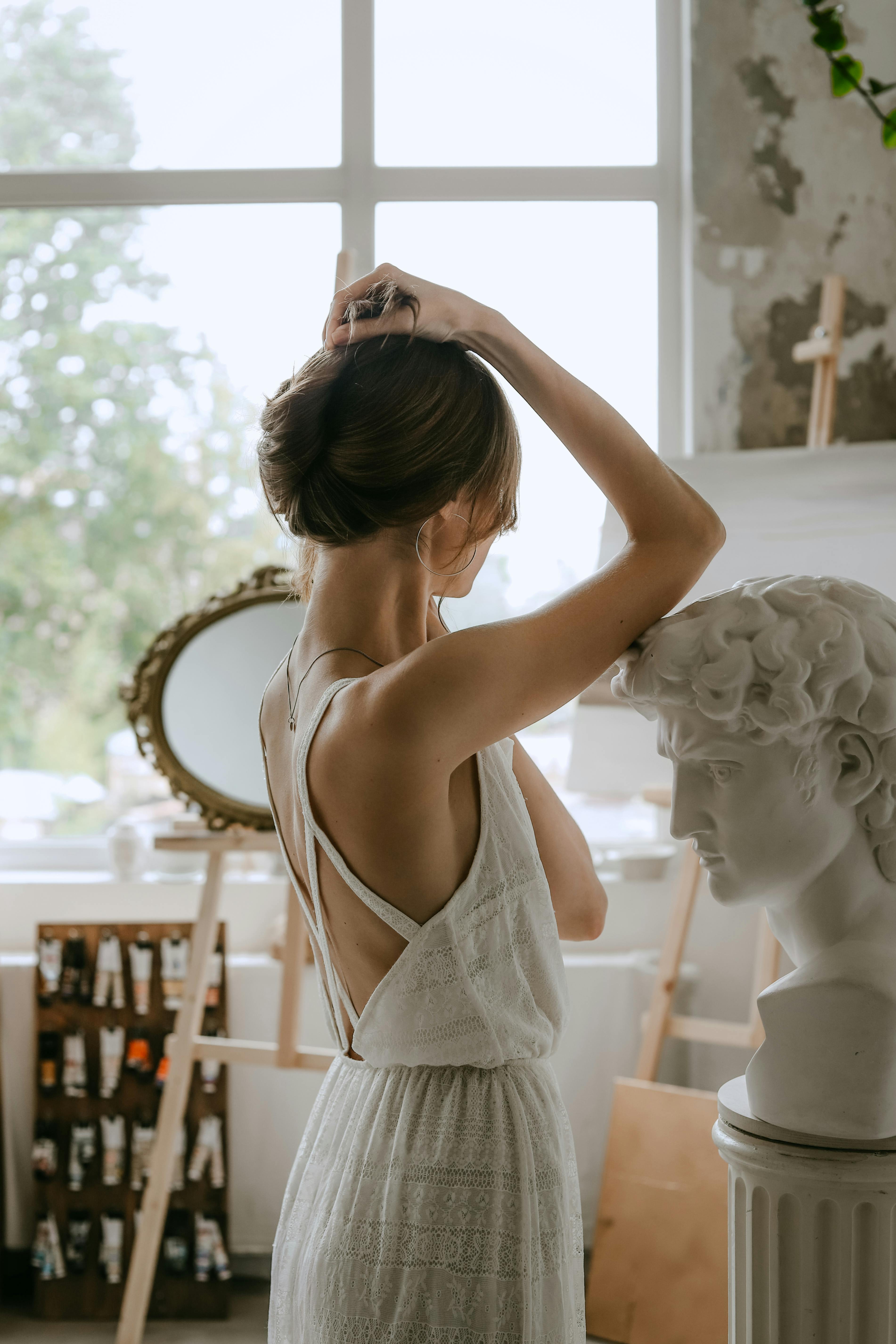 A woman in a white dress poses gracefully beside a classical bust sculpture in an artistic studio setting.