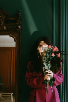 Fashionable woman in pink coat holding a bouquet of roses indoors with vintage decor.