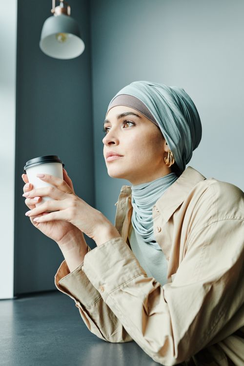 Free Young woman in hijab holding a coffee cup while gazing thoughtfully indoors. Stock Photo