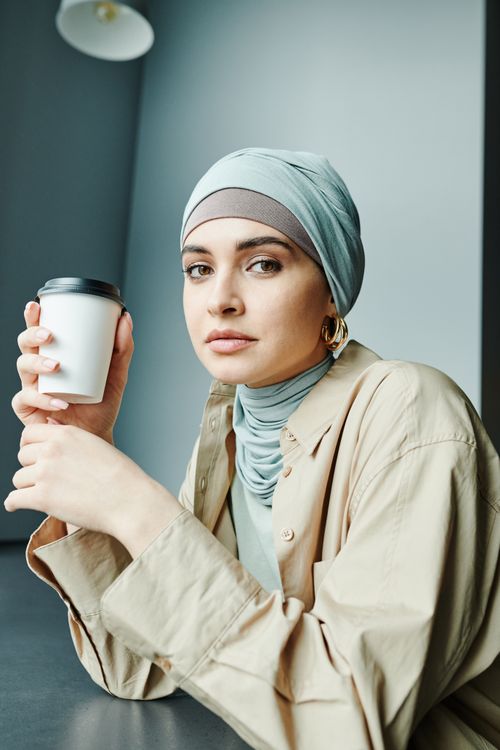 Free A stylish woman in a hijab holding a takeaway coffee cup indoors. Stock Photo