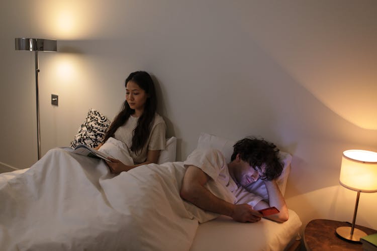 A Woman Sitting On The Bed Reading A Book Near The Man Lying Down While Using His Phone