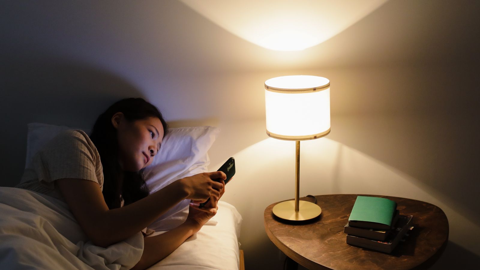 An adult using a smartphone in bed beside a lit table lamp