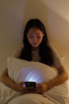 Asian woman smiling at phone while relaxing in bed during the night.