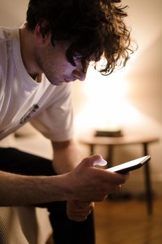 A young man in a cozy bedroom intently focused on his smartphone during evening hours.