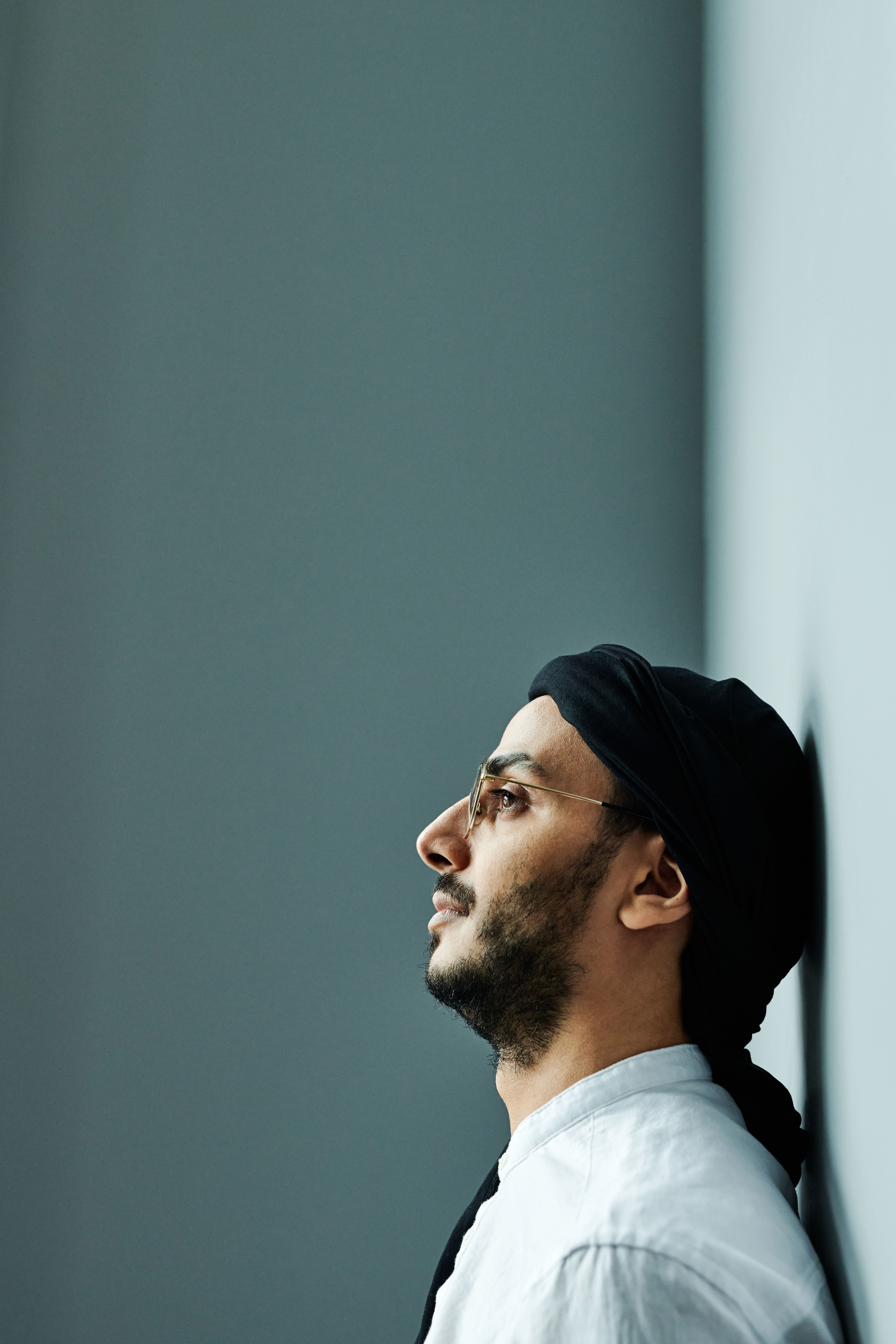 Side profile of a bearded man wearing a headscarf and glasses, leaning against a grey wall.