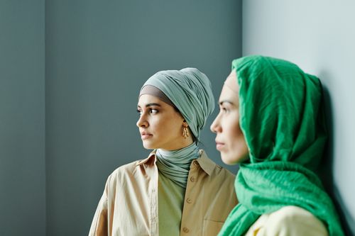 Free Two women in hijabs stand indoors, looking thoughtfully to the side in a serene setting. Stock Photo