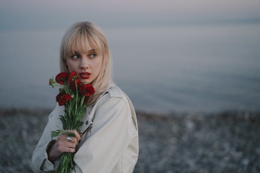 A young woman with red lips holds a bouquet of red flowers by the serene seaside.