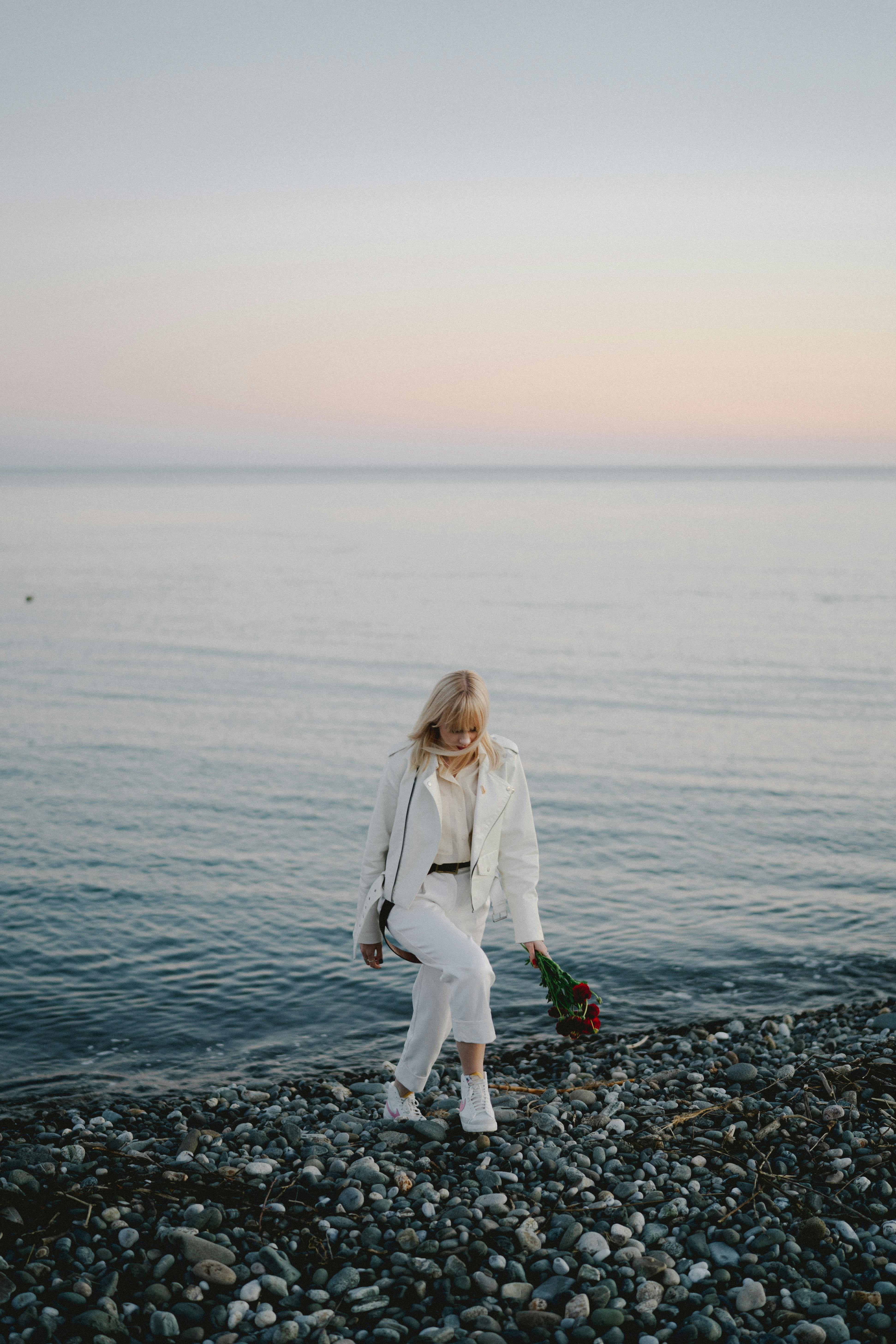 日落时分，一位白衣女子手持花束，沿着鹅卵石沙滩散步