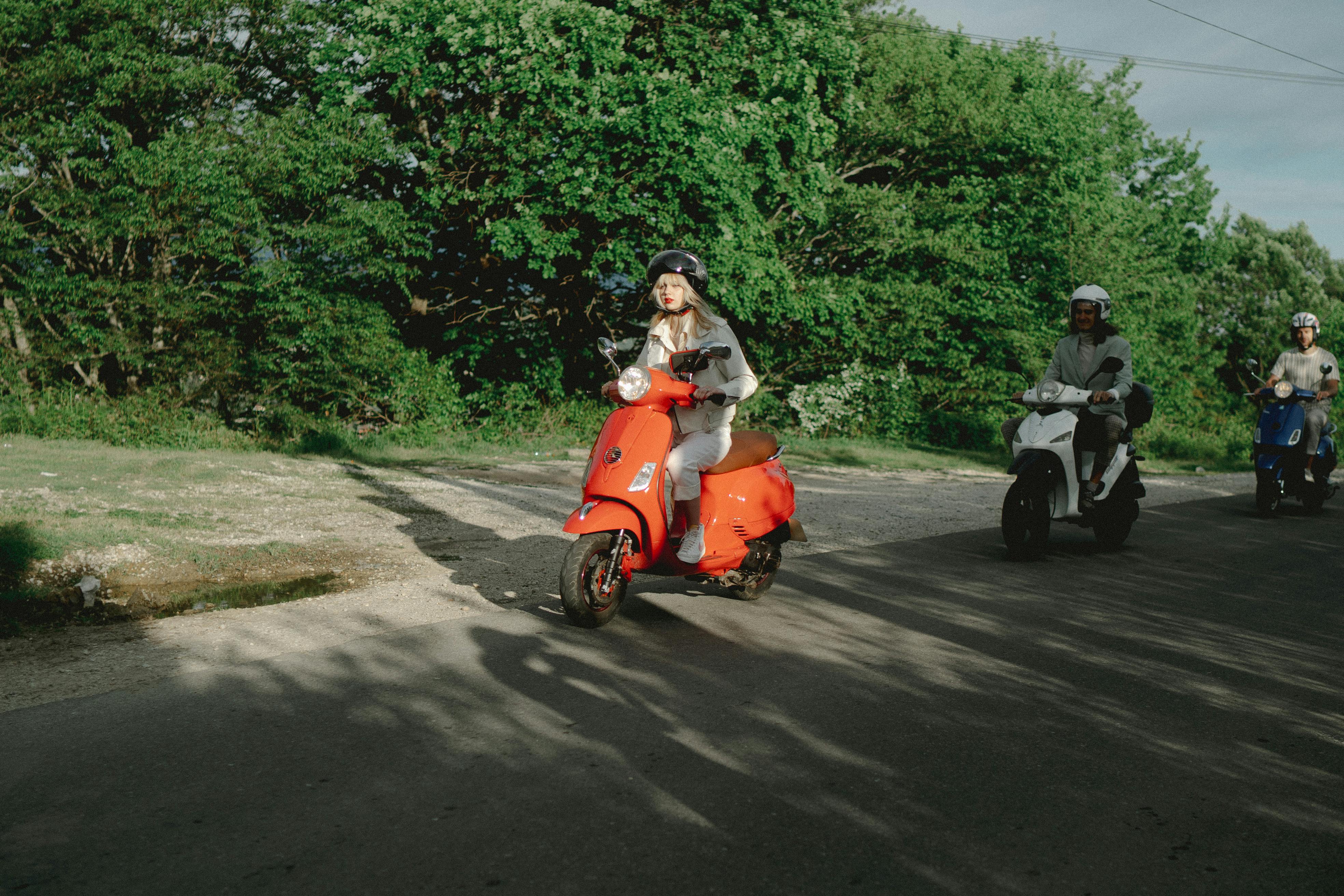 Three adults ride scooters on a sunny day, surrounded by lush greenery, enjoying the outdoors.