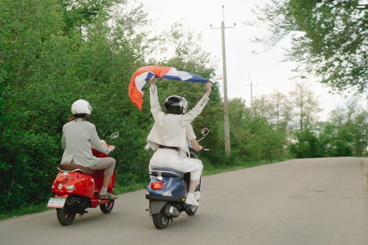 People Riding A Motor Scooters On The Road While Holding A Flag