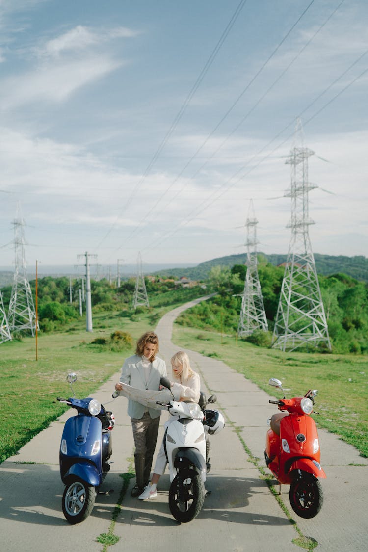 A Man And Woman Looking At The Map On The Road While Standing Near The Motorcycles