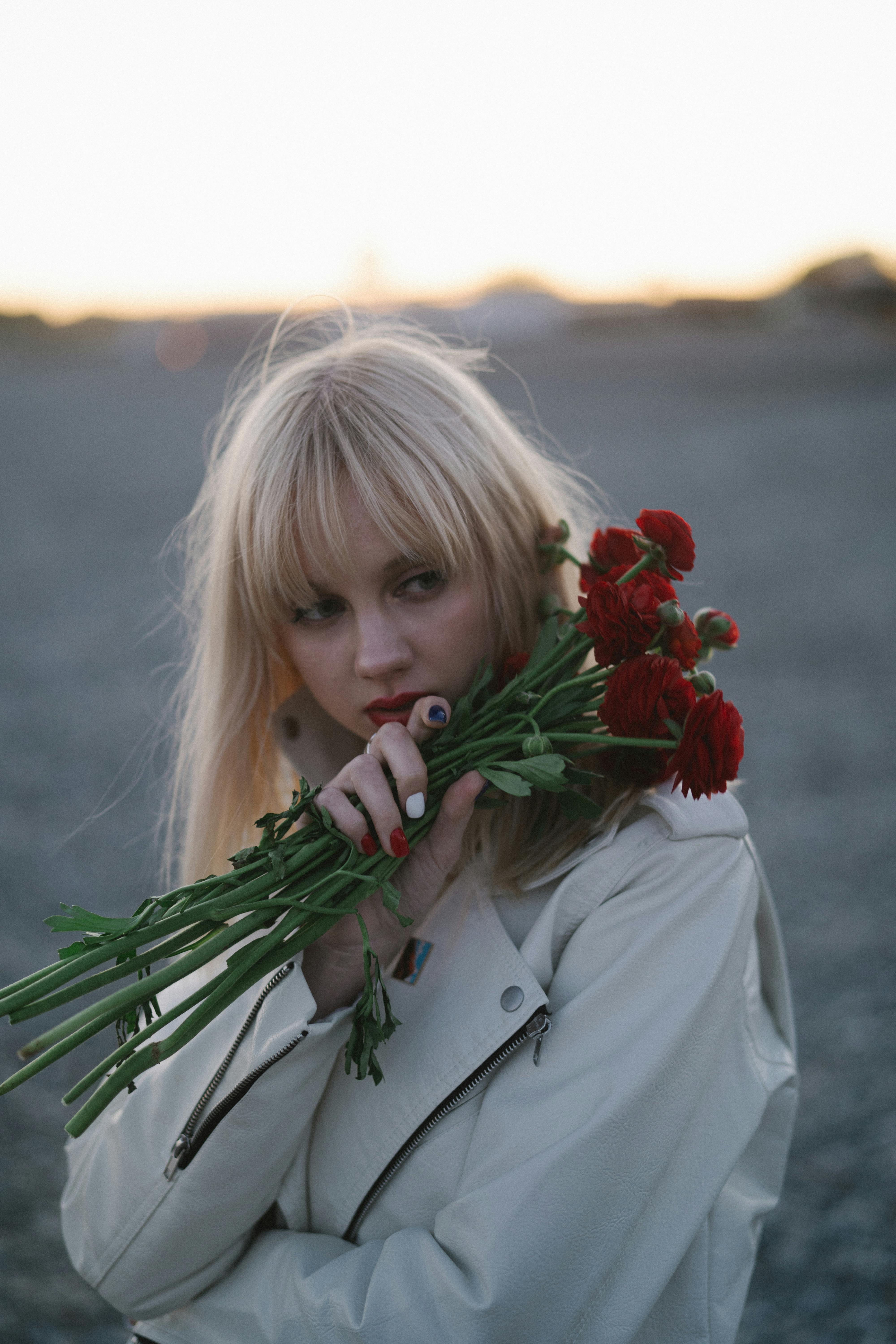 Blonde woman in white coat holding red flowers at sunset outdoors.