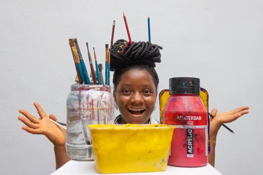 Smiling artist with dreadlocks surrounded by painting materials including acrylics and brushes.