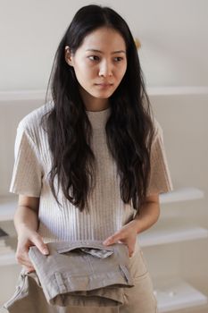 An adult woman in a beige shirt holds folded clothes, appearing thoughtful in a minimalist indoor setting.