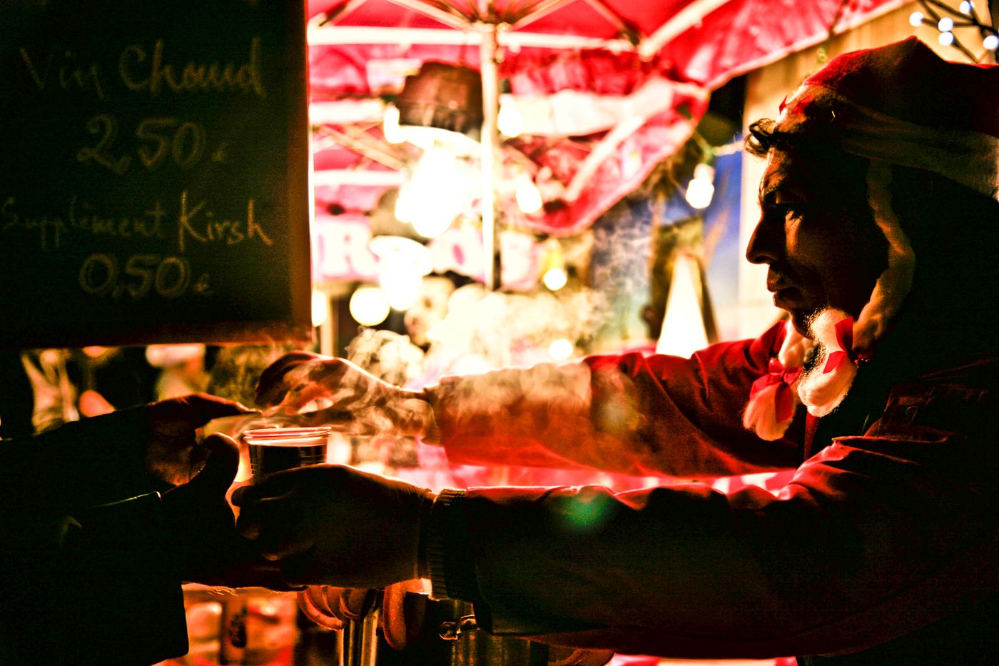A man in a Santa hat serves hot wine at a Christmas market in Lyon, France at night.