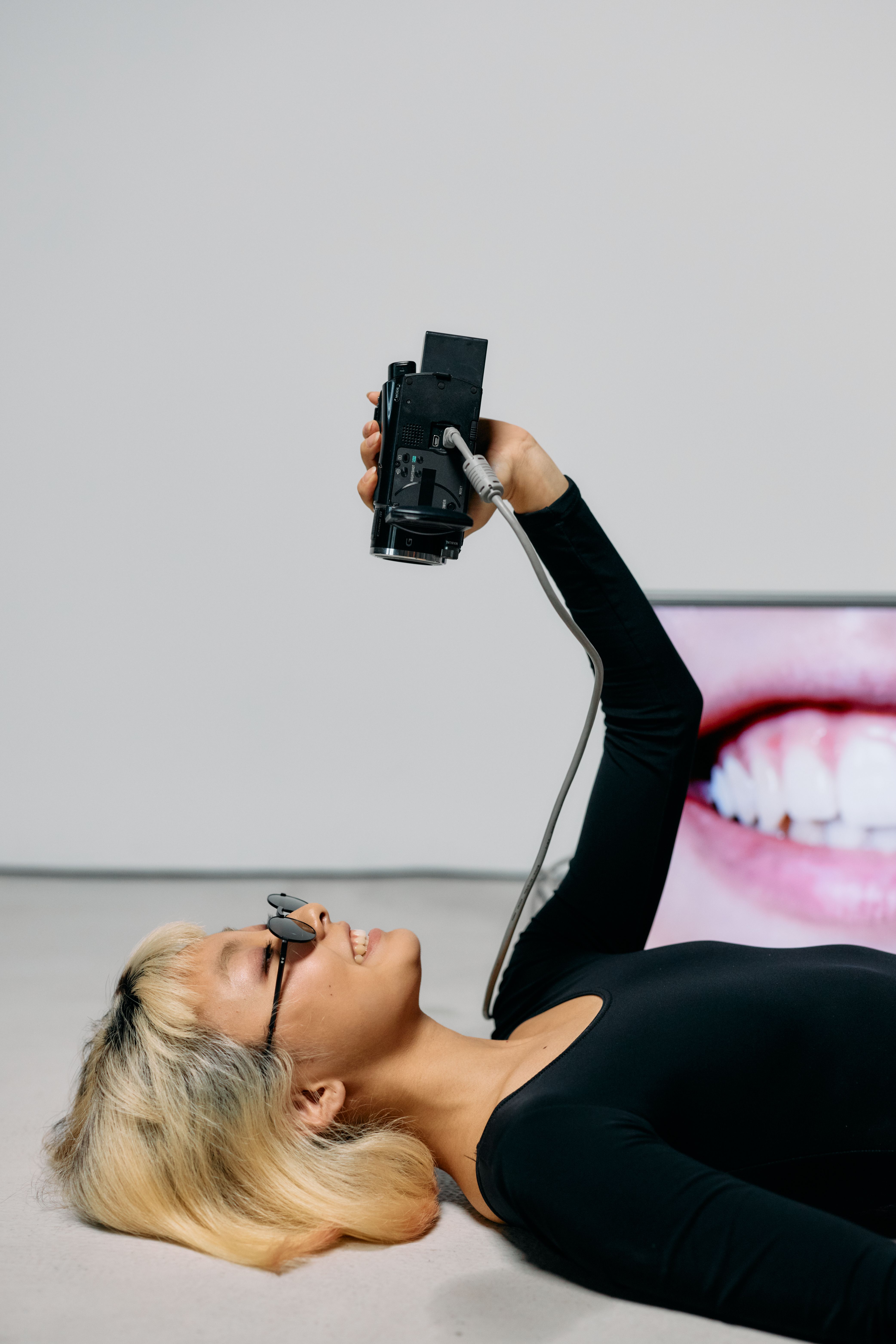 Trendy fashion model with sunglasses lying down using a video camera in a modern studio setting.