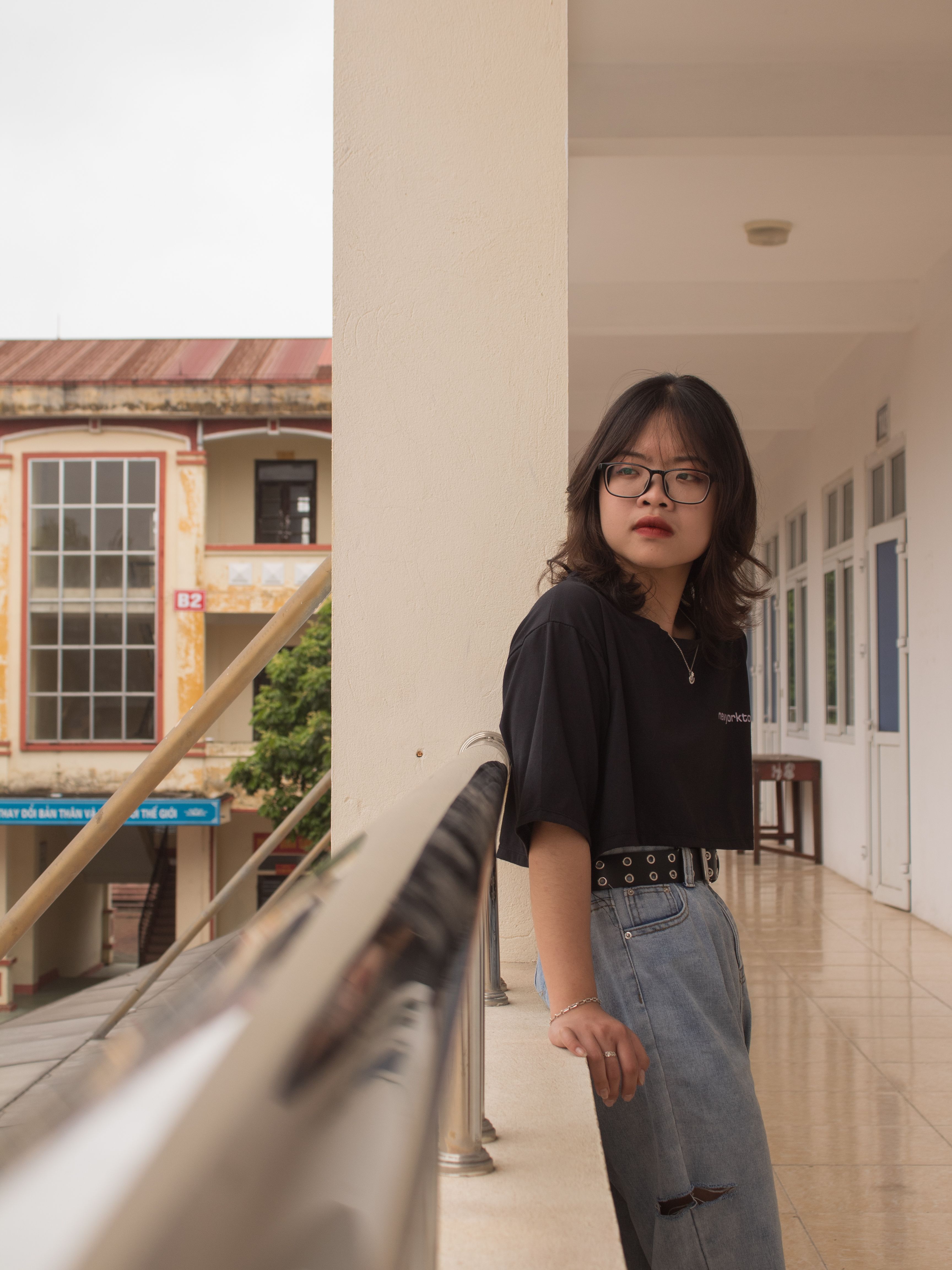 A young woman in casual attire leans on a balcony railing in a Vietnamese city.