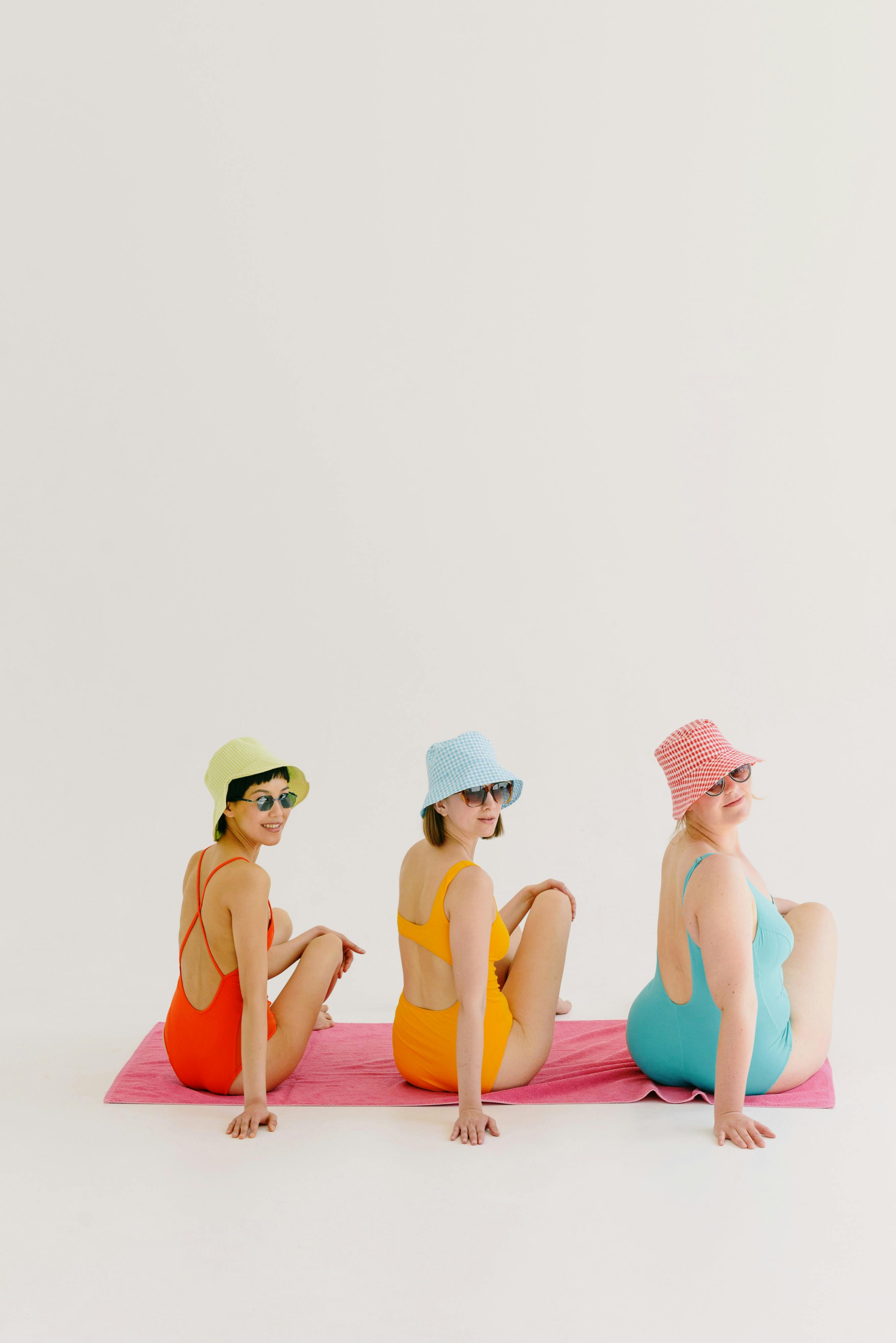 Three young women in colorful swimsuits posing sitting on a towel with bucket hats and sunglasses.