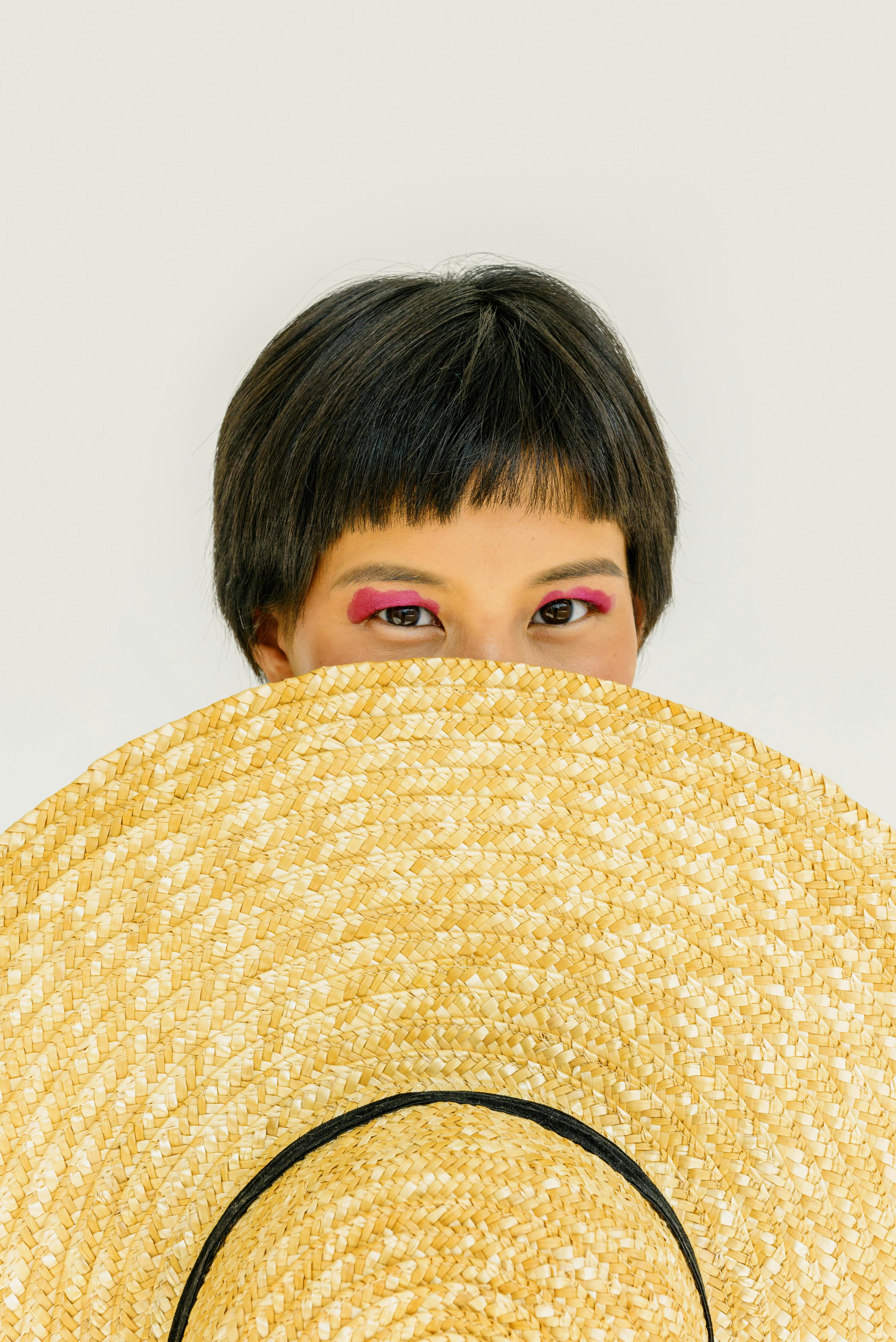 A woman with short hair and eye makeup peeks over a large straw hat in a studio shoot.