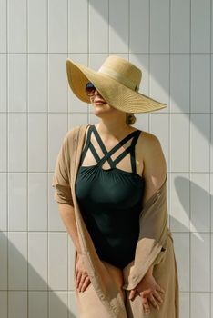 Fashionable adult woman in swimwear with a sun hat poses against a tiled wall under bright sunlight.