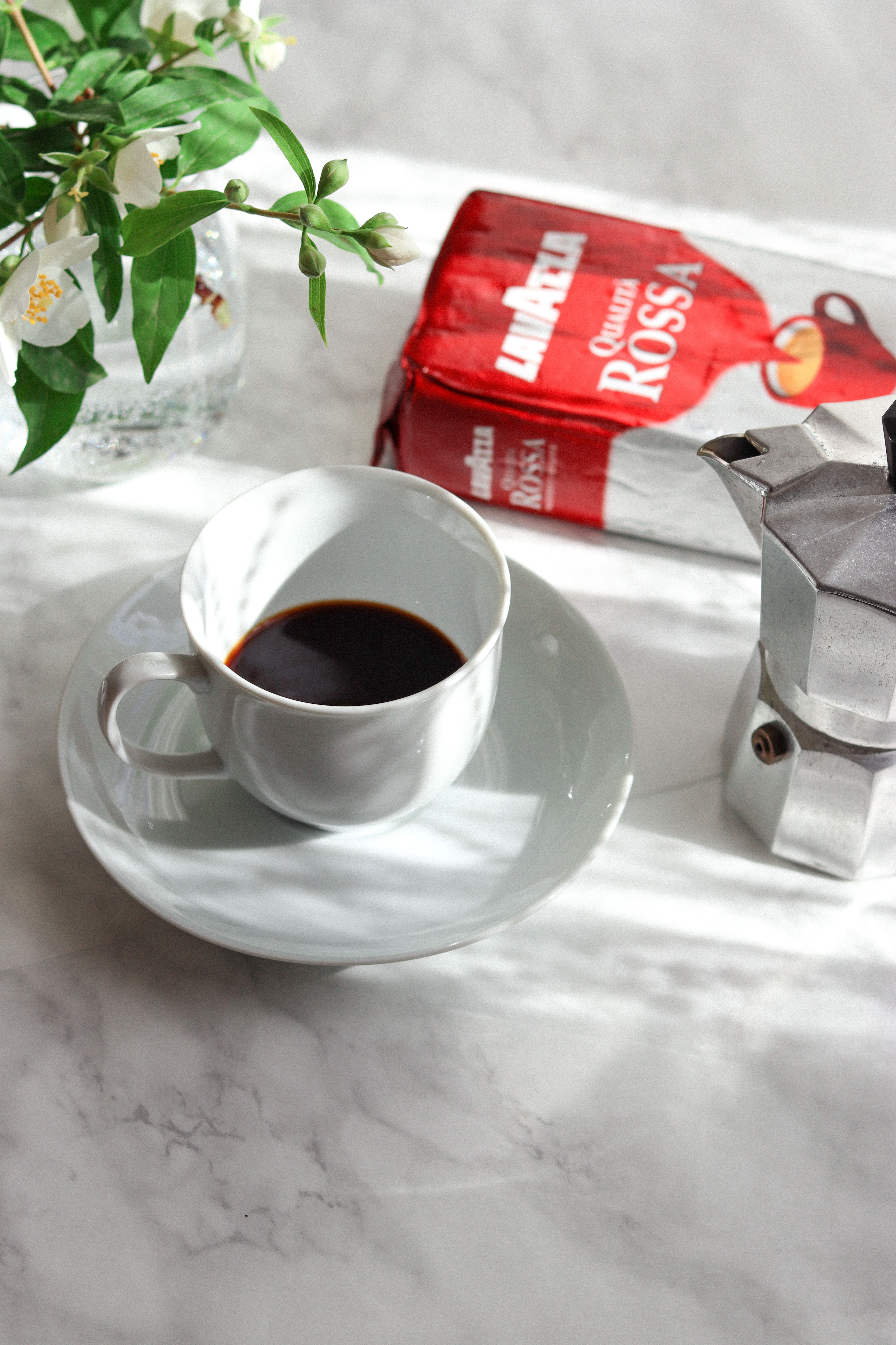 An elegant coffee setting featuring a white cup of coffee, Italian coffee package, and a stovetop kettle.