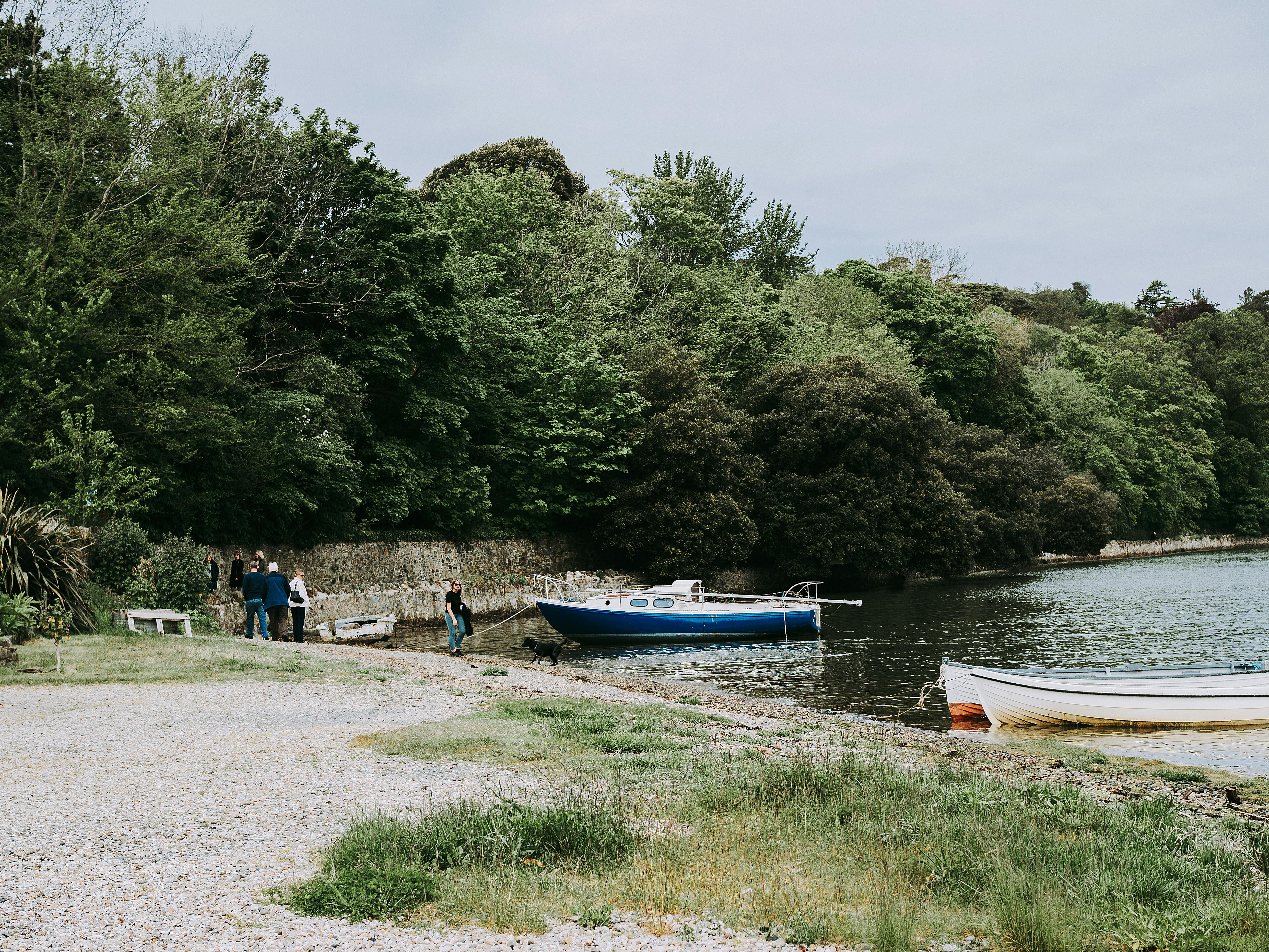 Tranquil lakeside with boats docked, surrounded by lush green trees and a pebble beach.