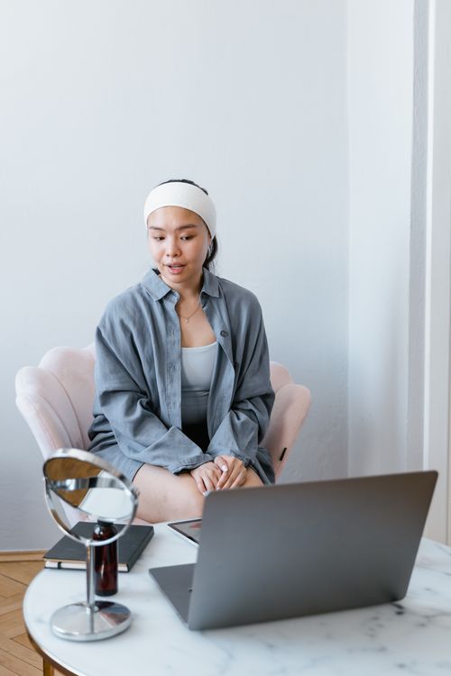 Free Woman sitting indoors, engaged with laptop and mirror, fostering a wellbeing routine. Stock Photo