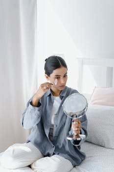 Woman sitting on bed using a jade roller for skincare, wearing casual clothing in a bright room.