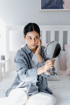 A woman sitting on a bed, gazing into a handheld mirror in a serene bedroom.