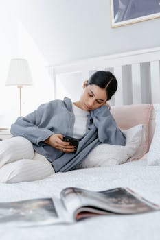 Woman in pajamas sitting on bed using smartphone in bright airy bedroom.