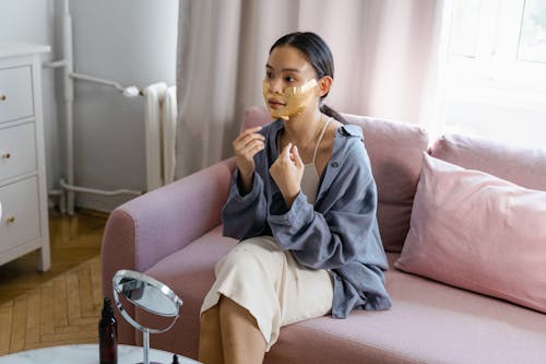 Young Asian female with facial mask applied on face sitting on couch near table with mirror during skincare routine at home