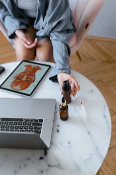 Top-down view of skincare products and jade roller on a table near a laptop and a tablet showing skincare routine.