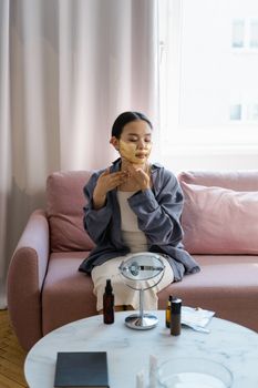 Asian woman applying gold facial mask at home, sitting on a cozy sofa with a skincare routine.