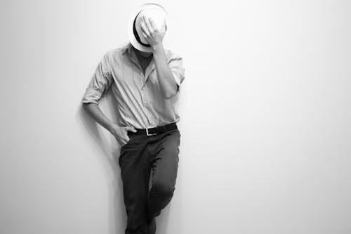 Grayscale Photo of Man Leaning on Wall Wearing a Fedora Hat