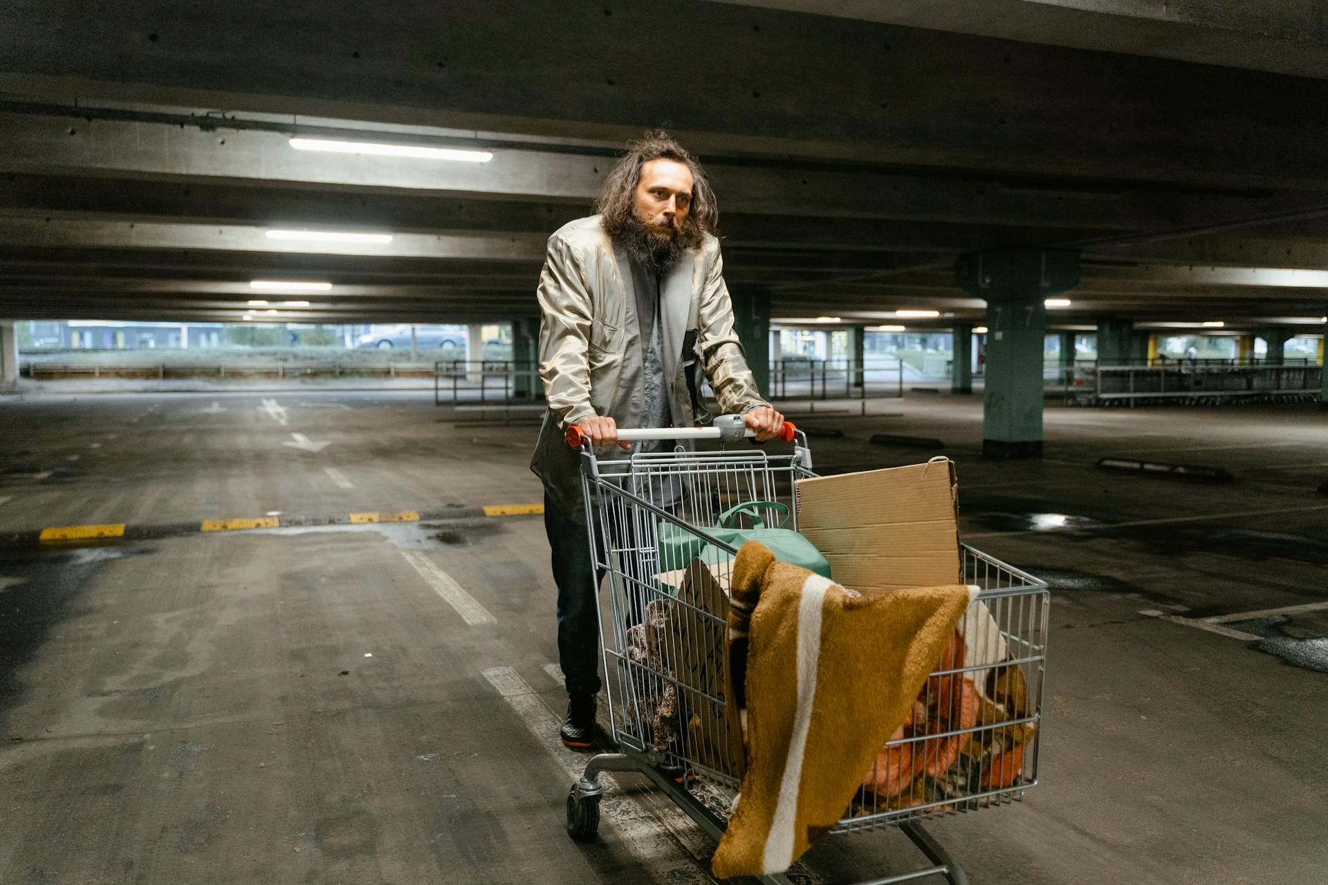 Adult male pushing a shopping cart filled with belongings in an empty parking garage.