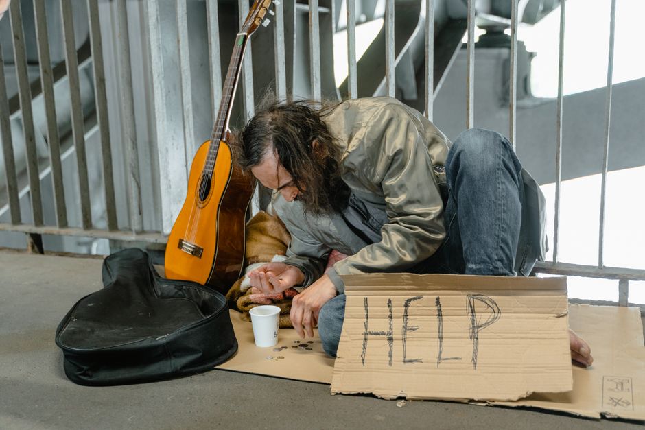 Un hombre sin hogar está sentado junto a una barandilla con un cartel de cartón que dice 'AYUDA', monedas y una guitarra.