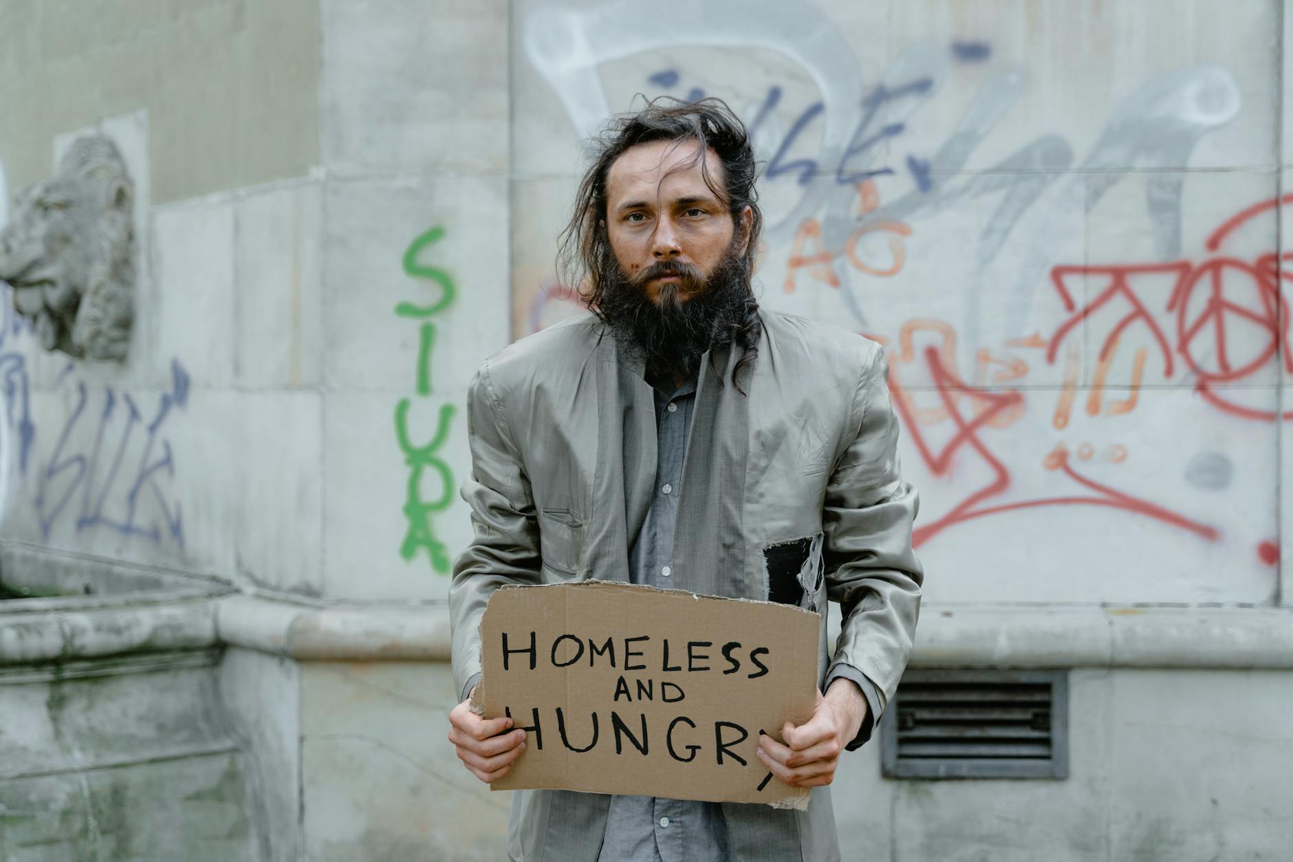 Homeless man standing on the street holding a sign, with graffiti in the background.