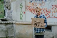 A homeless woman standing outdoors holding a sign that reads 'Will Work For Food'.